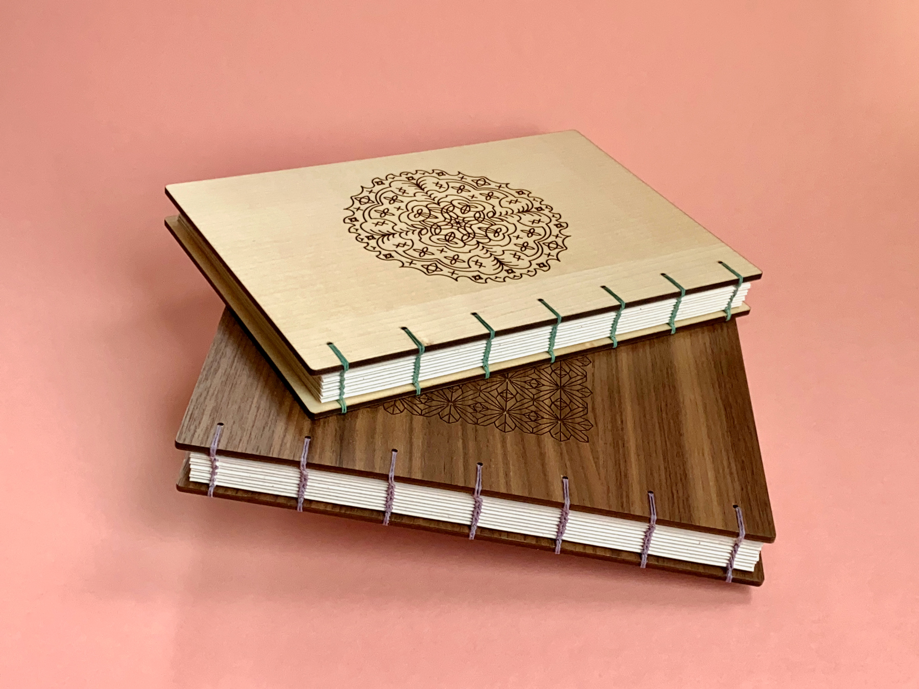 Two coptic-bound books one maple, one walnut lay on top of each other against a rose pink background.